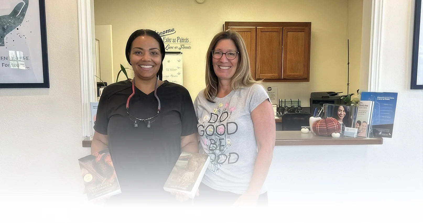 Two women smiling and holding books at a dental office reception counter.